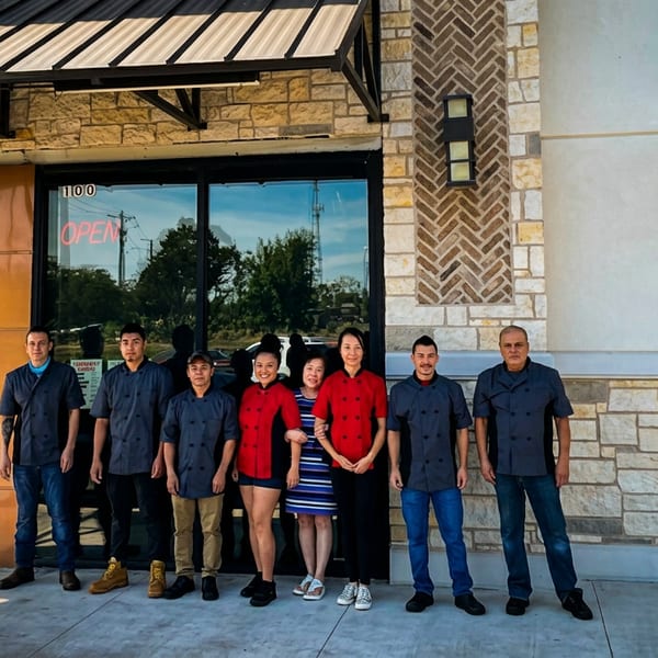 Seven restaurant staff in chef coats and one woman stand outside the restaurant entrance.