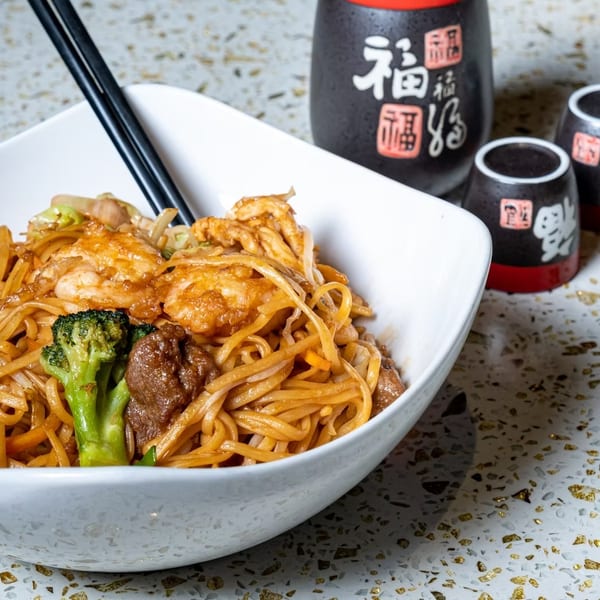 Bowl of stir-fried noodles with shrimp, beef, vegetables, and chopsticks, accompanied by black-and-red Japanese sake cups.