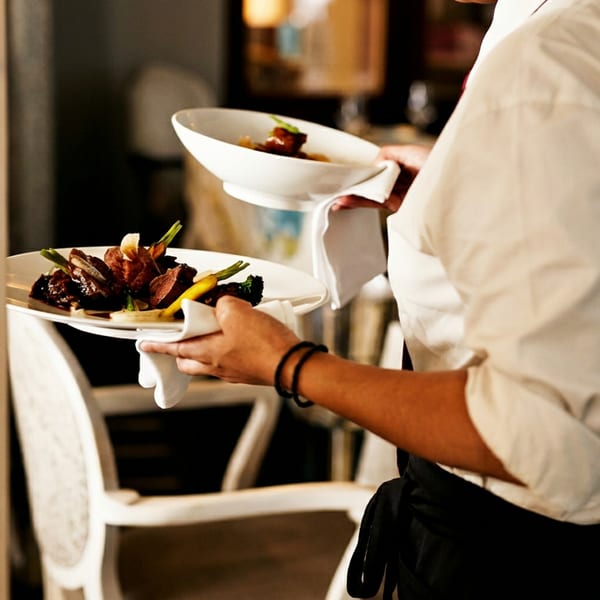 Server in a white shirt and black apron carries two plates of beautifully plated food.